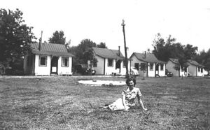 Black-and-white photograph of a woman sitting on grass in front of a row of cabins